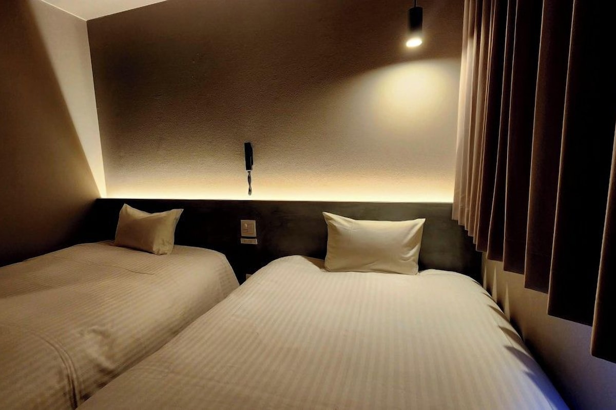 Une chambre d’hôtel avec deux lits, une tête de lit dotée d’un luminaire et une fenêtre avec des rideaux.