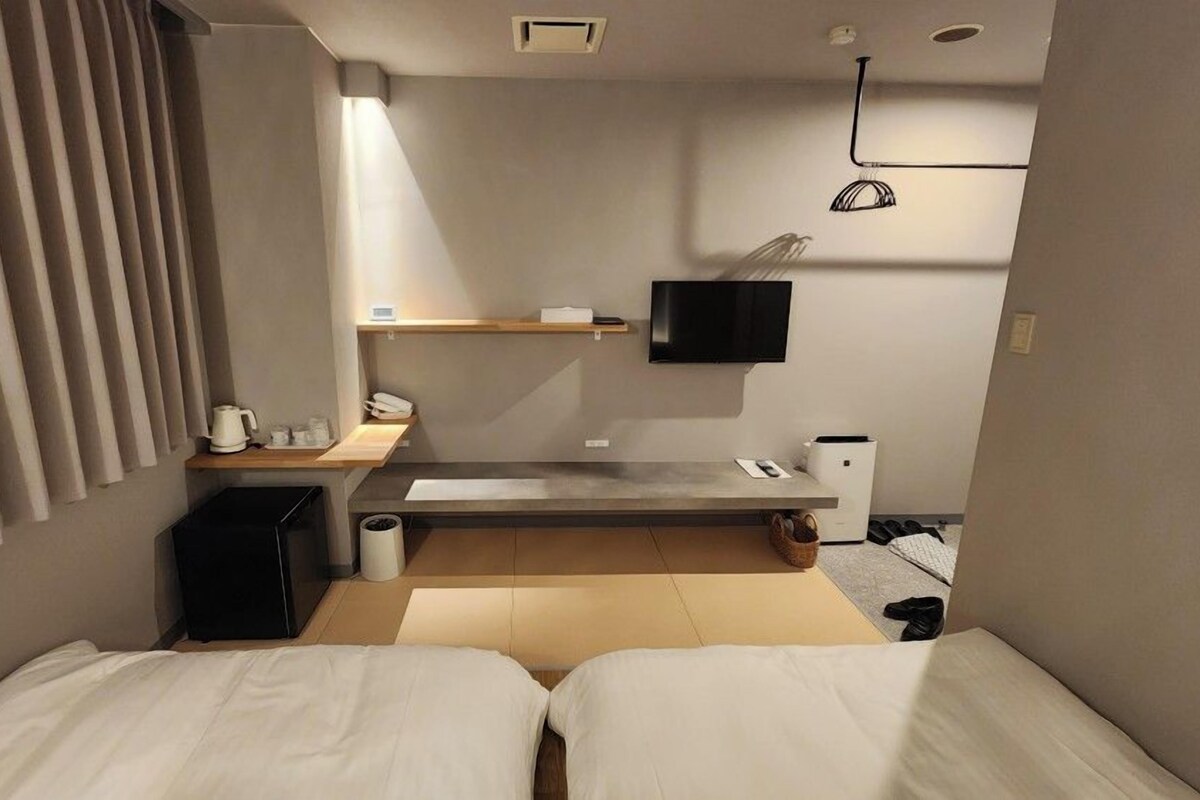 A compact hotel room with two single beds, a small desk, a television, and a kitchenette area.