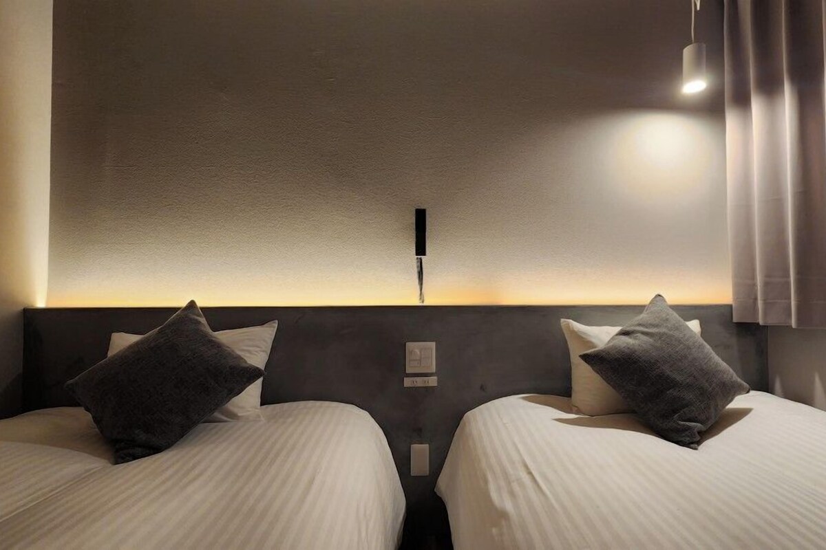 A hotel room with two beds, a nightstand, a wall-mounted light, and a window with curtains.