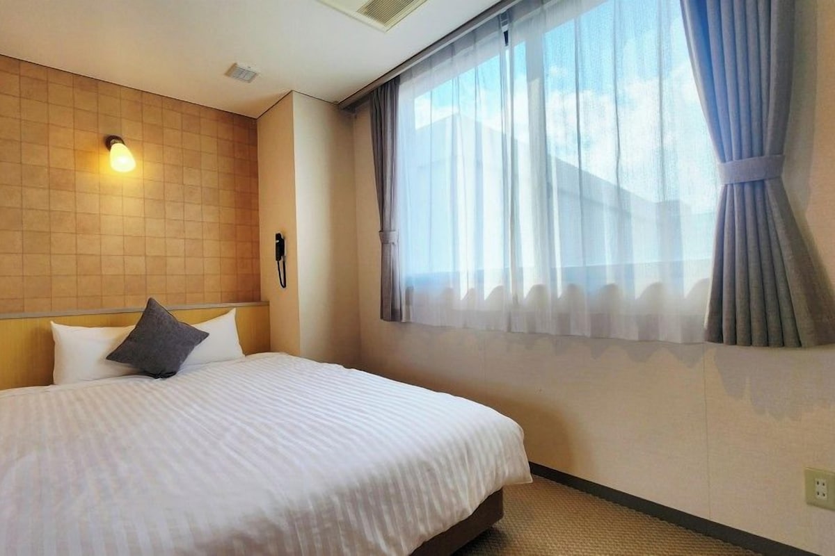 Une chambre d’hôtel avec un lit, une tête de lit, un téléphone fixé au mur et une fenêtre avec des rideaux.
