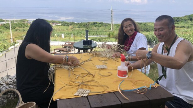 Tres personas participan en una actividad en una mesa al aire libre con vista al mar y vegetación de fondo.