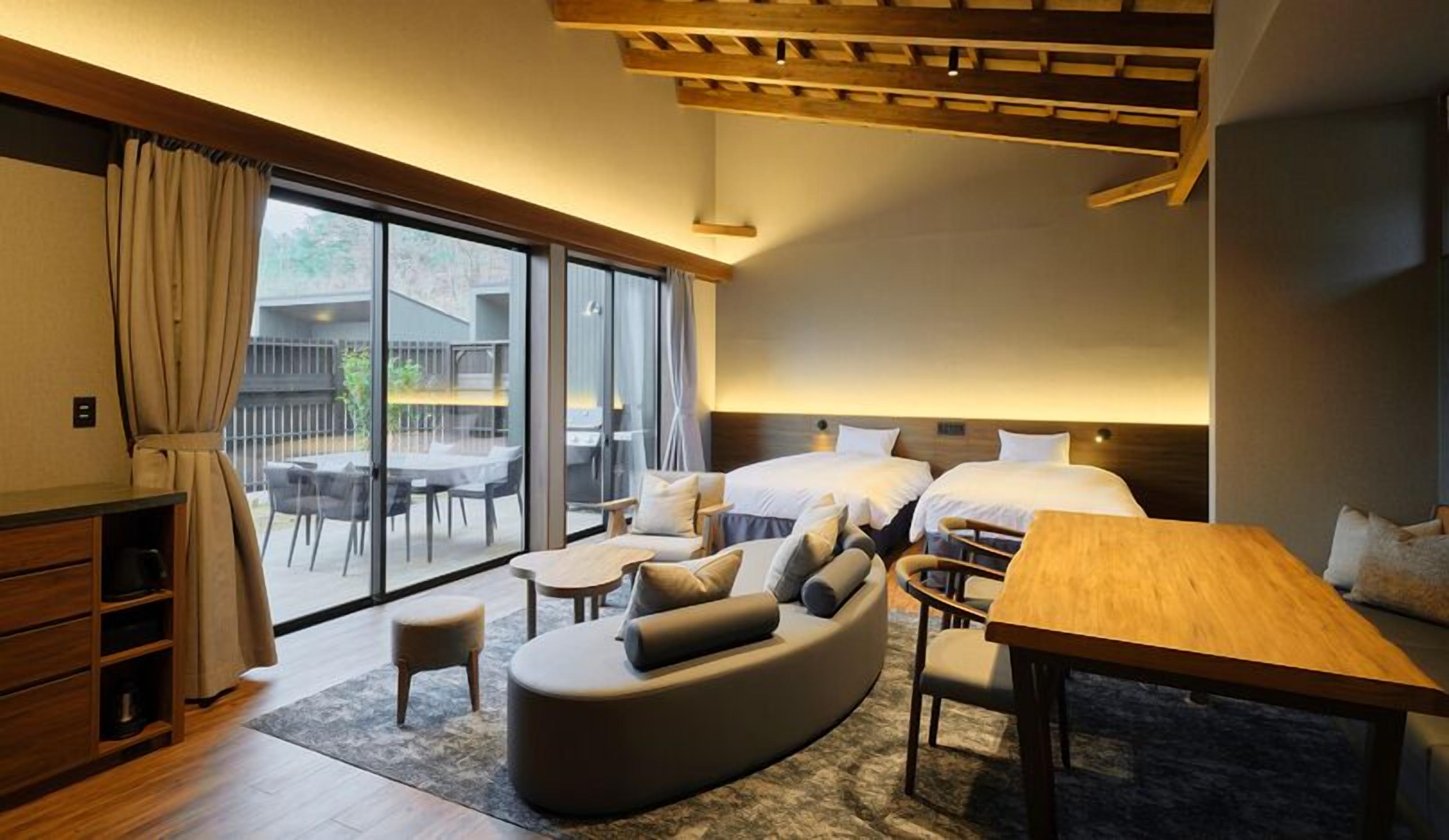 A modern hotel room with a sofa, two beds, a dining table, and a balcony with outdoor seating.