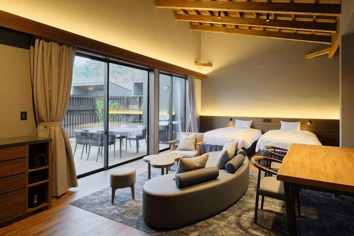 A modern hotel room with a sofa, two beds, a dining table, and a balcony with outdoor seating.