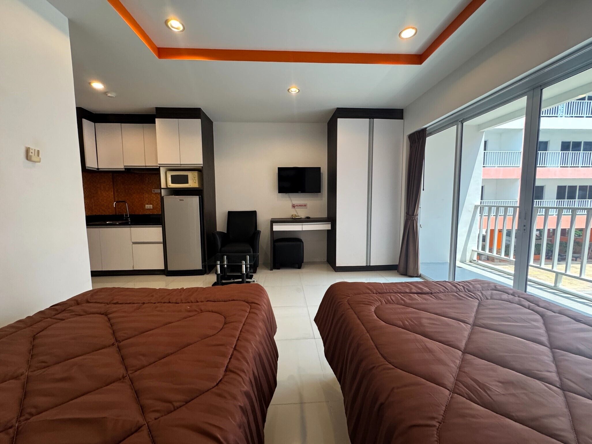 A hotel room with two beds, a kitchenette, a TV, and a balcony with a view of a building.