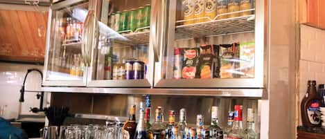 A bar area with a refrigerator stocked with beverages, a selection of liquor bottles, and a counter with glasses and bottles.