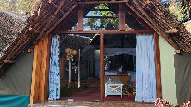 A traditional wooden structure with a thatched roof, a glass door entrance, and a small table inside. - The giraffes cottages restaurant and bar (Nairobi)
