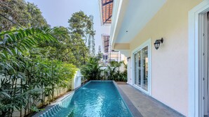 A rectangular swimming pool with a tiled edge, surrounded by lush greenery and a building with a light-colored exterior and a balcony with a wooden roof.
