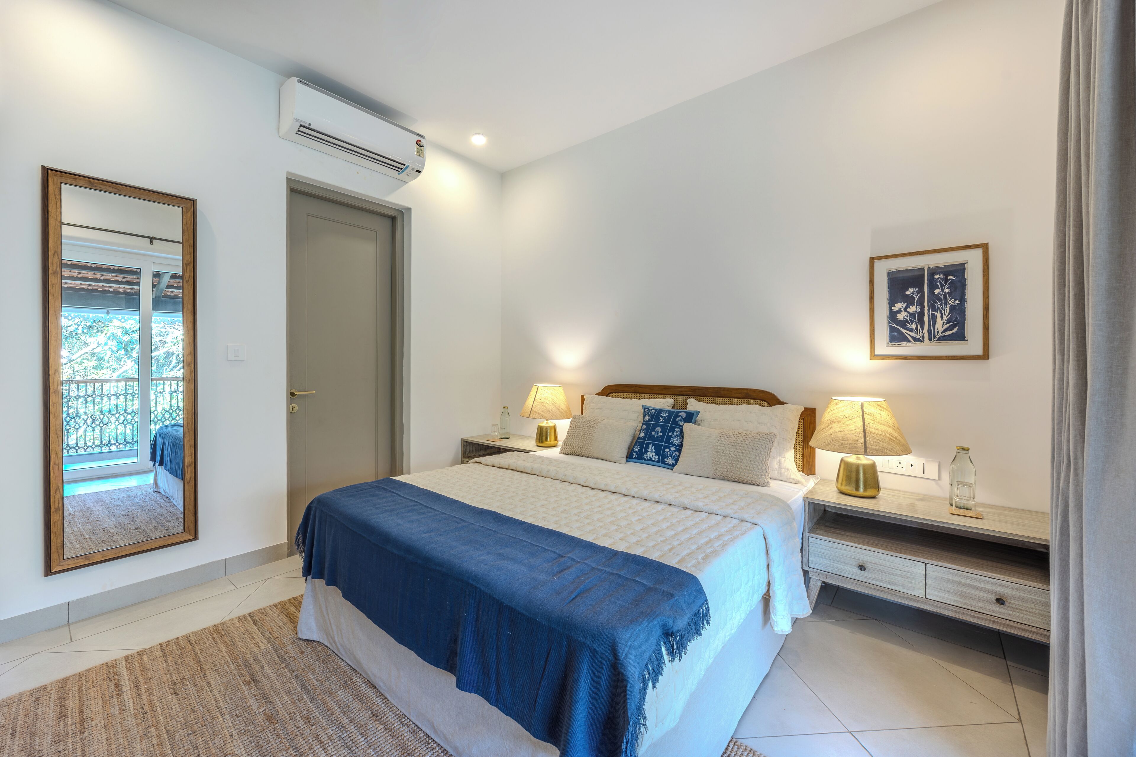 A bedroom with a bed, bedside tables, a mirror, an air conditioner, and a window with a view.