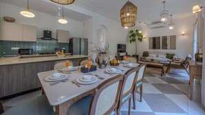 A modern kitchen with a dining area, featuring a large island, hanging lights, and a view of the living room.