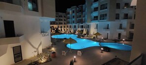 Ein Nachtblick auf einen Hotelinnenhof mit mehreren Pools, Liegestühlen und Sonnenschirmen.