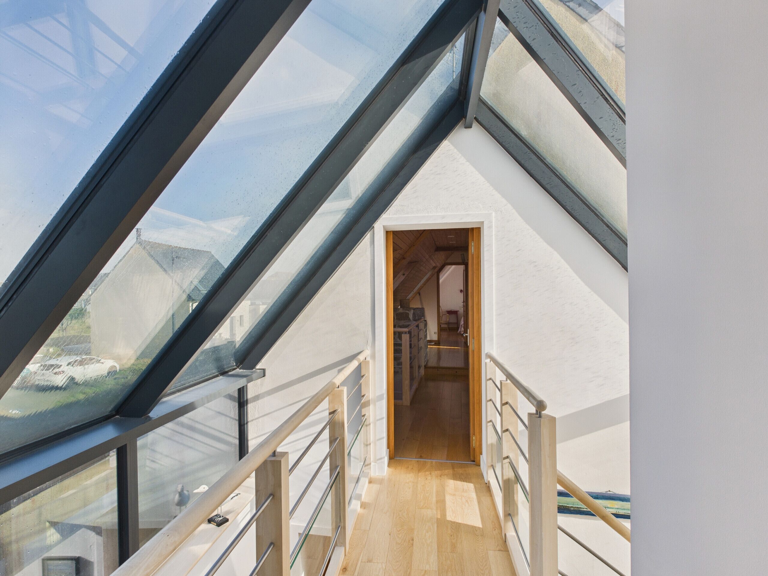 A modern interior with a sloped glass roof, wooden flooring, and a view through a doorway.