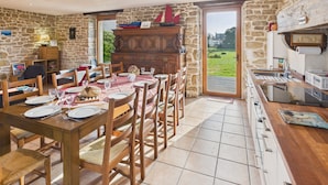 A dining area with a wooden table set for four, surrounded by chairs. A stone wall and a wooden cabinet with a sailboat model are visible. - Beautiful house for 8 with garden – Sea view (Penmarch)