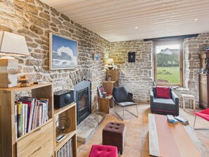 A cozy living room with a stone wall, a fireplace, a bookshelf, a sofa, and a view of the outdoors. - Beautiful house for 8 with garden – Sea view (Penmarch)