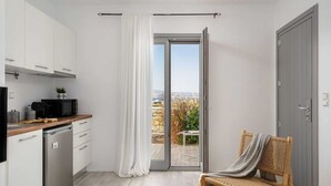 A compact kitchen with a sliding glass door leading to a balcony, a wicker chair, and a grey door. - Julies, Sunset Bliss Apartment – Agia Anna (Naxos)