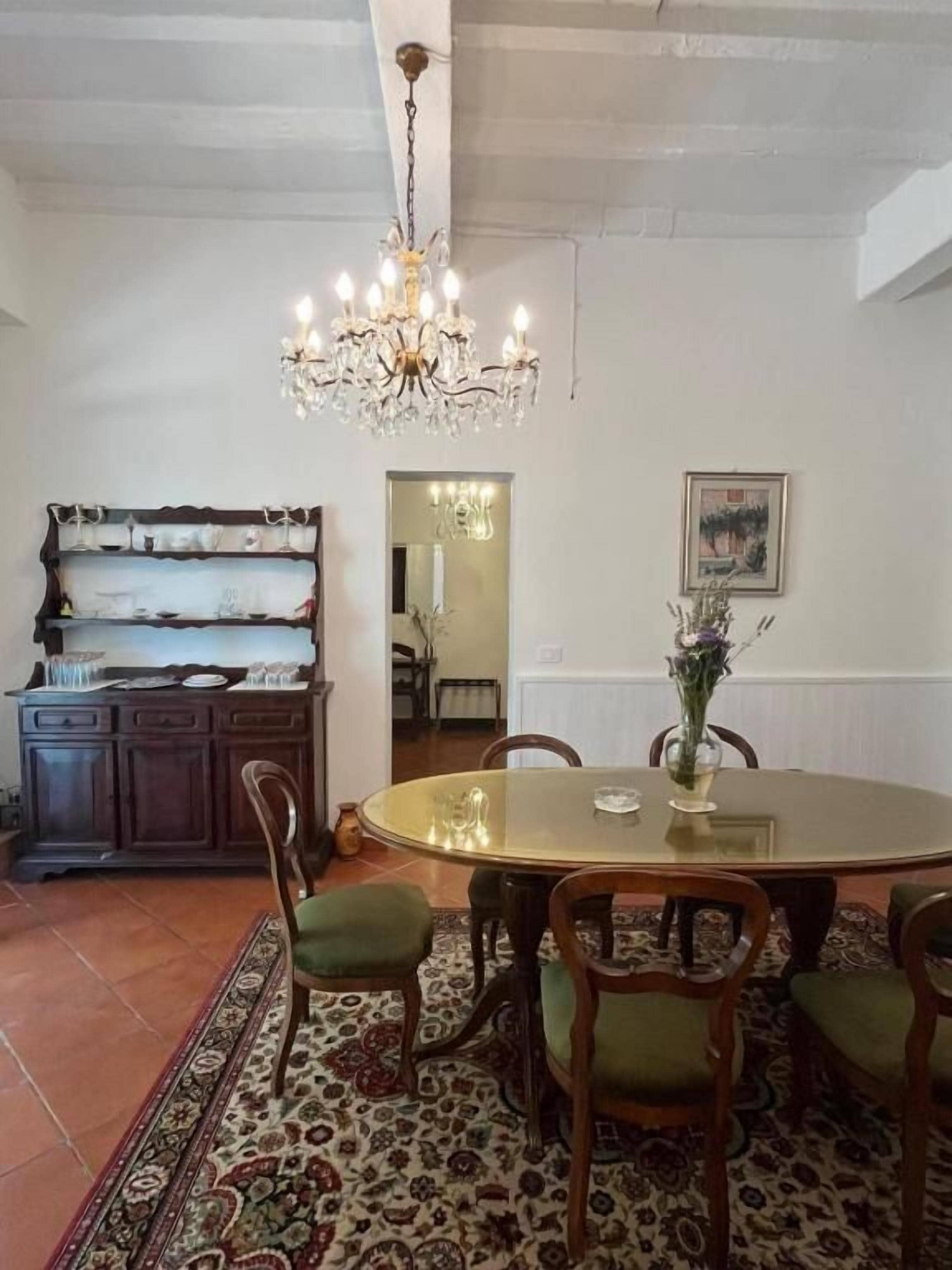 A dining area with a round glass table, six wooden chairs with green cushions, a chandelier, a wooden buffet with glassware, and a patterned rug.