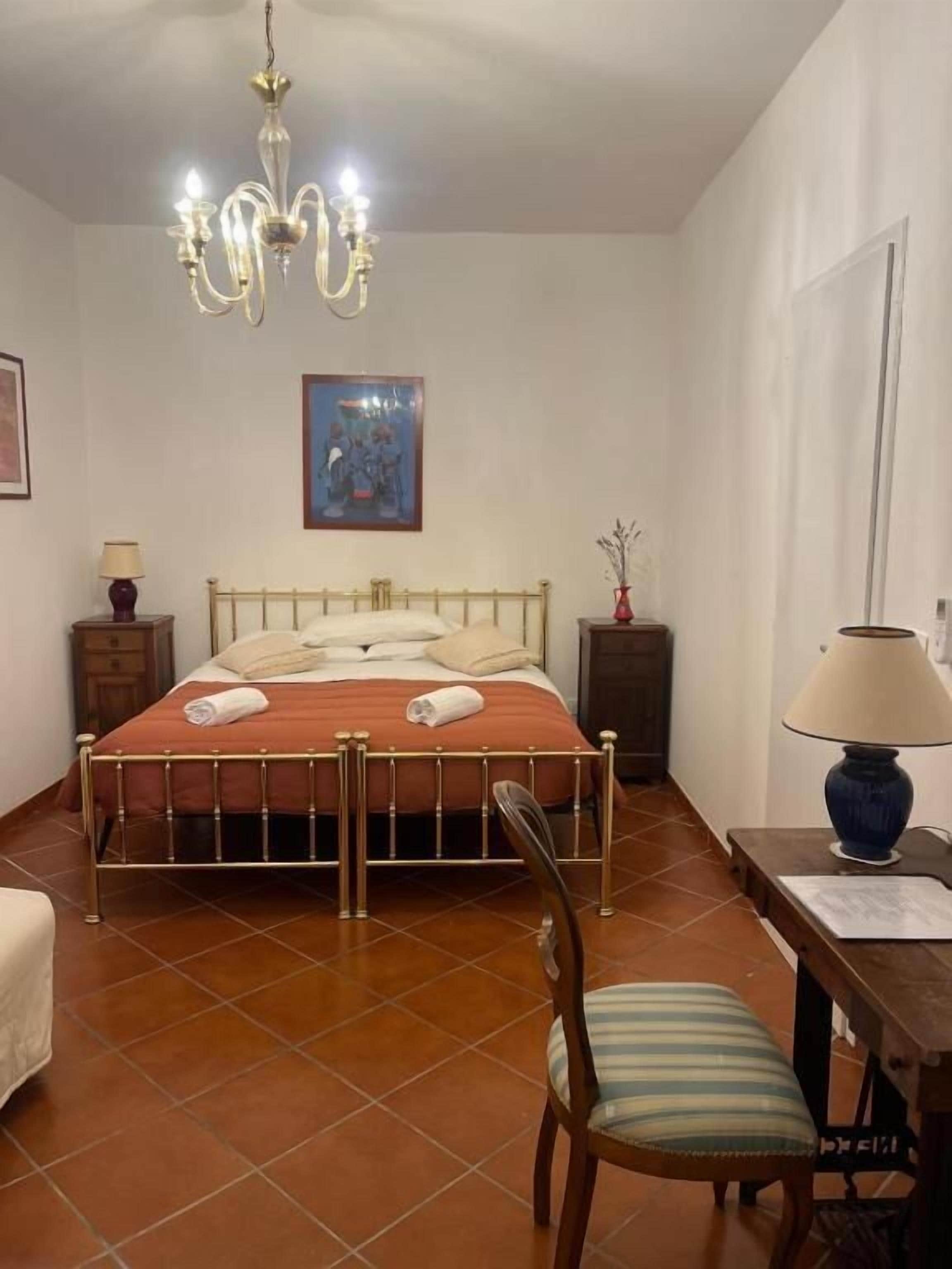 A bedroom with a bed, a chair, a desk, a lamp, a painting, and a chandelier.