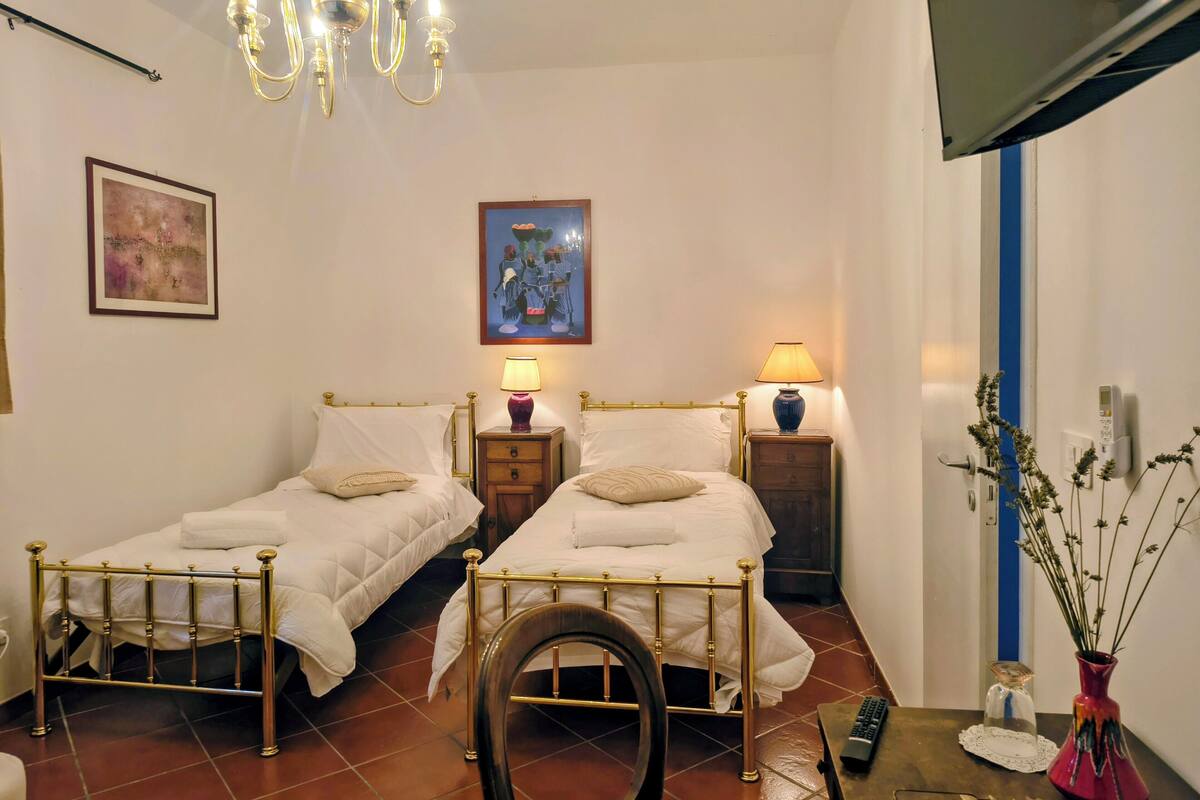 Quarto de hotel com duas camas de solteiro, um candelabro, uma pintura, um abajur, um vaso com flores e uma televisão fixada na parede.