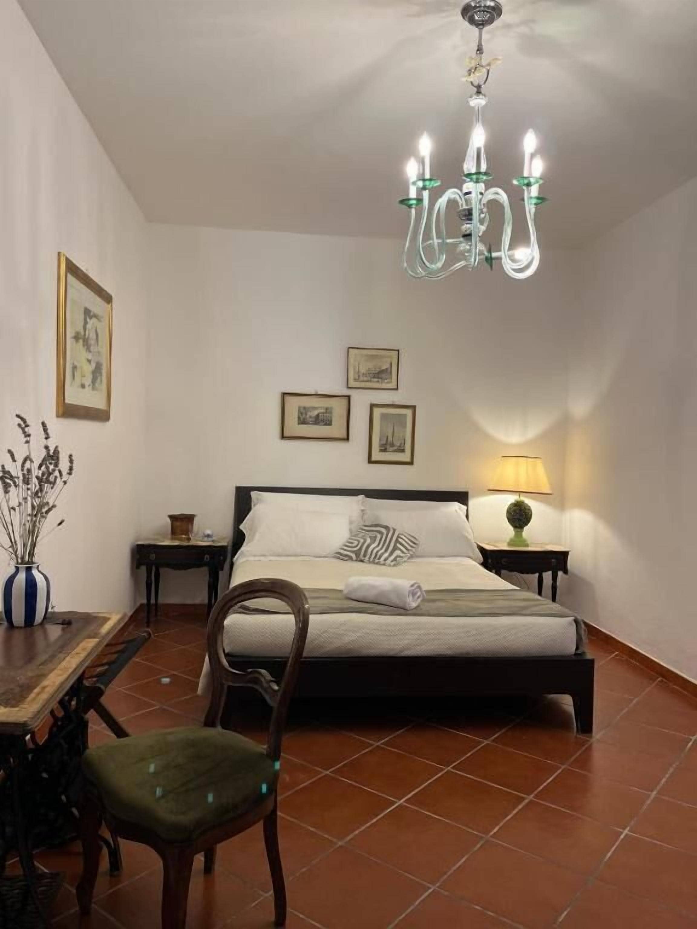 A bedroom with a bed, a chair, a table, a lamp, and a chandelier.