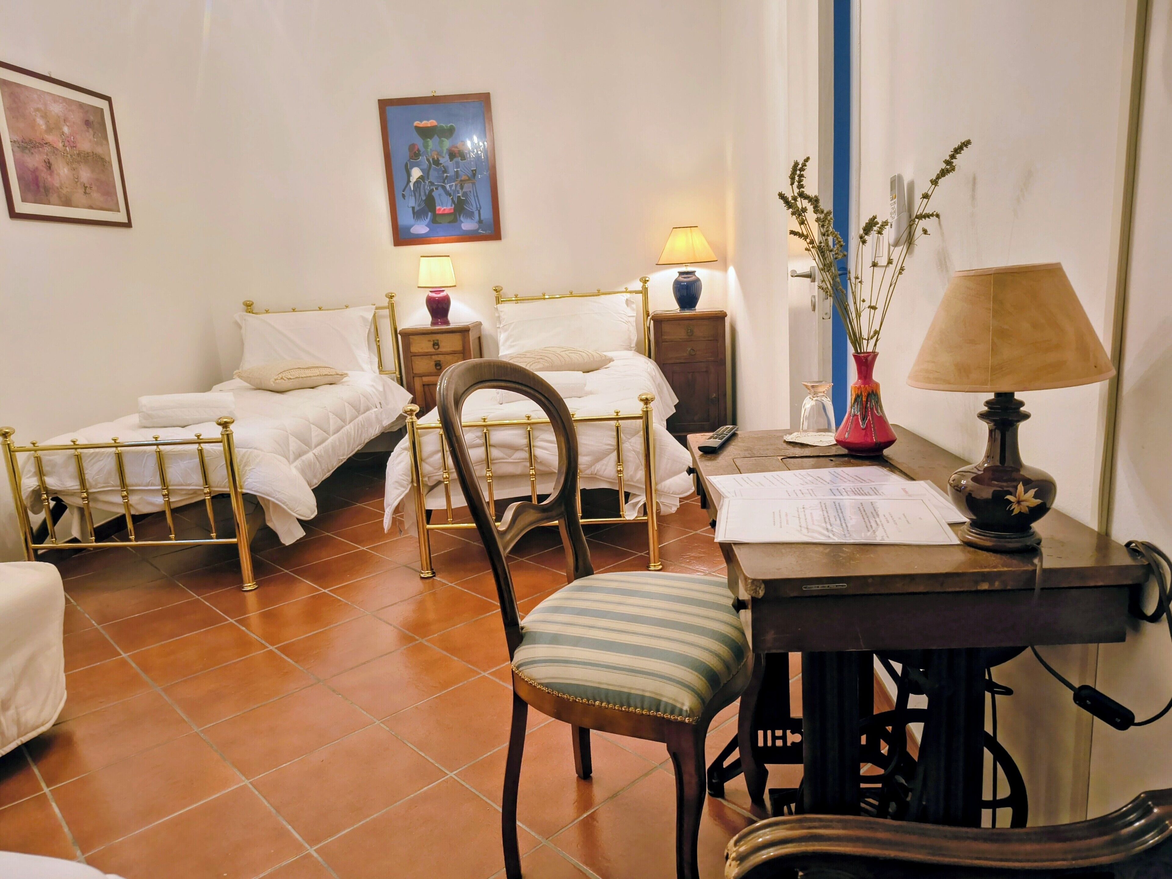 A room with two single beds, a wooden chair, a desk with papers, a lamp, and a vase with plants.