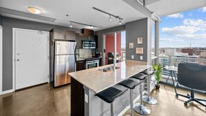 A modern kitchen with a stainless steel refrigerator, microwave, and stove, a bar area with stools, and a large window offering a city view. - DT Views | UG Park | Walk to Saddledome |AC (Calgary)