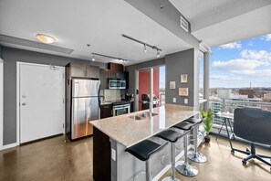A modern kitchen with a stainless steel refrigerator, microwave, and stove, a bar area with stools, and a large window offering a city view. - DT Views | UG Park | Walk to Saddledome |AC (Calgary)