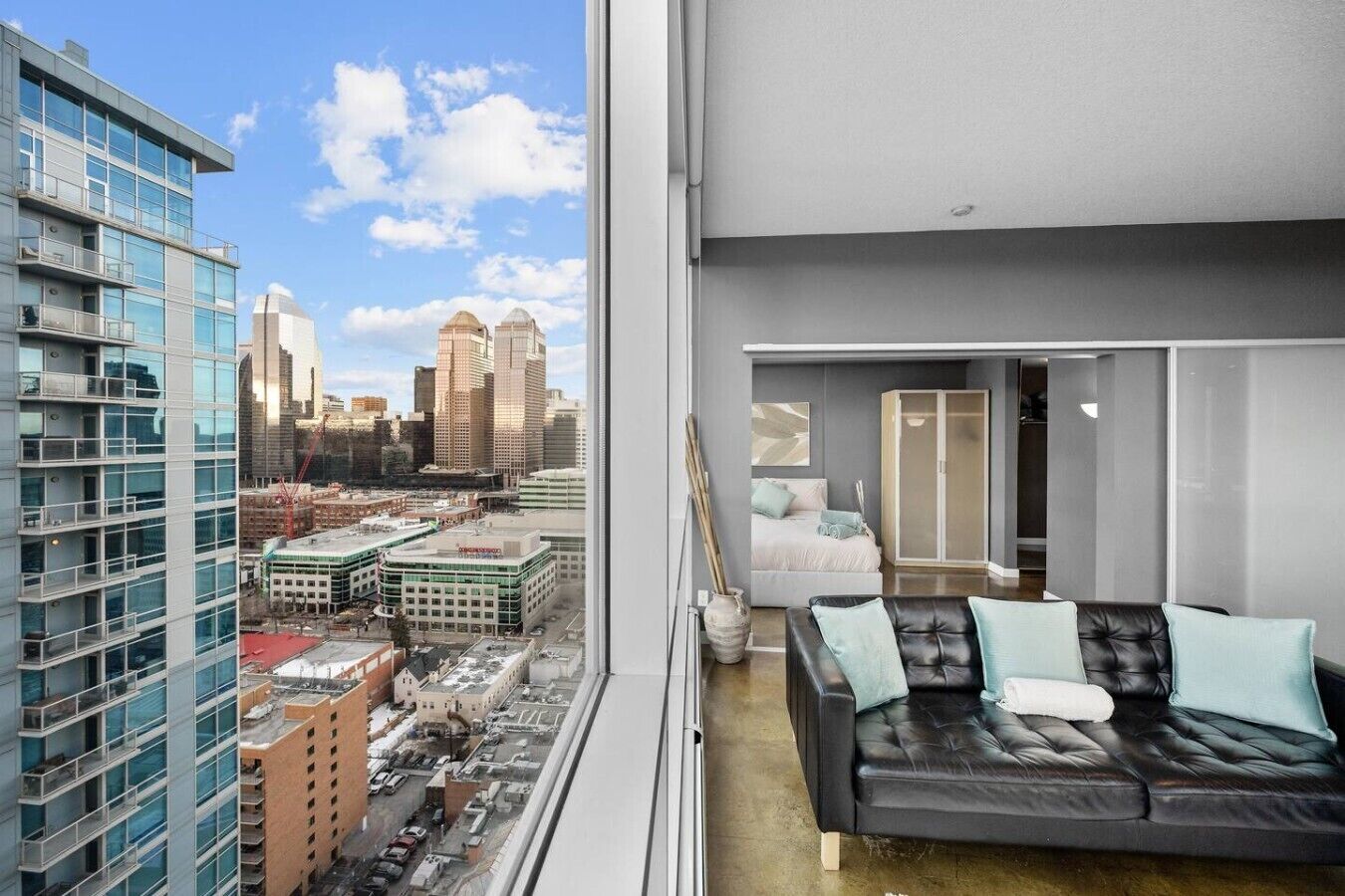 A modern interior with a black leather sofa, a bed with a white headboard, and a view of a cityscape through large windows.