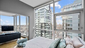 A modern bedroom with a large bed, a sofa, and a TV, featuring large windows and a city view. - DT Views | UG Park | Walk to Saddledome |AC (Calgary)
