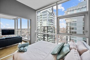A modern bedroom with a large bed, a sofa, and a TV, featuring large windows and a city view. - DT Views | UG Park | Walk to Saddledome |AC (Calgary)