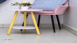 A small table with a white top and yellow legs, a pink chair with a blue cushion, and a potted plant with yellow flowers. - Maison Recla (Lomé)