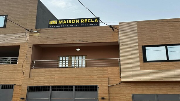 A building with a sign reading "MAISON RECLA" and a contact number. It has two large garage doors, a balcony with a railing, and a window with blinds. - Maison Recla (Lomé)