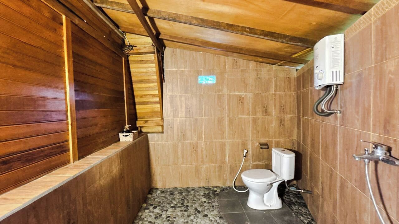 A small, wooden-walled bathroom with a toilet, a shower, and a water heater mounted on the wall.
