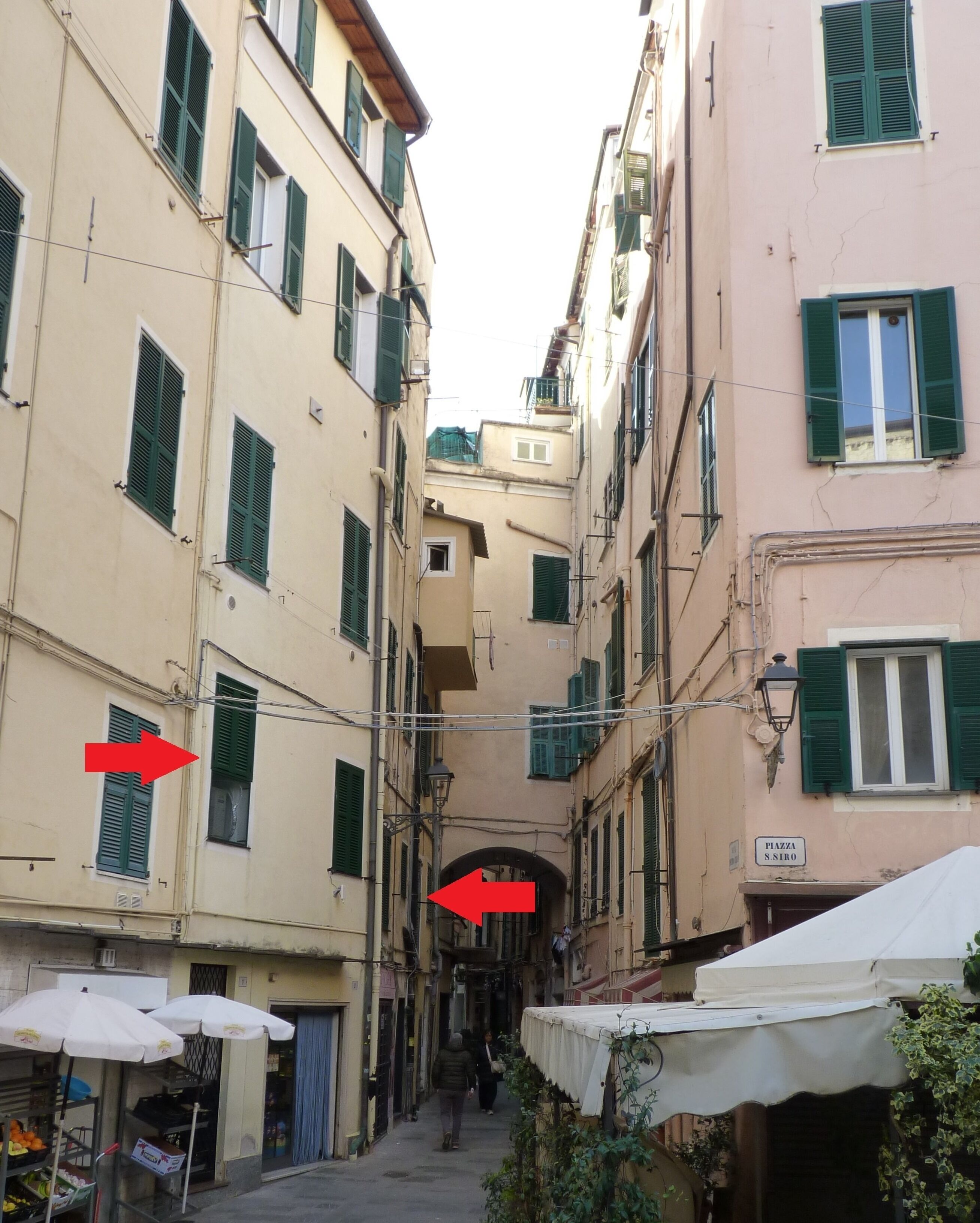 A narrow street with buildings on either side, featuring balconies and green shutters.