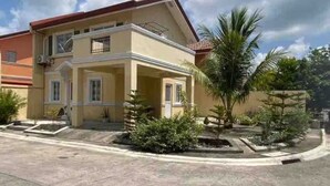 Una casa de dos pisos con un balcón, un pequeño jardín con palmeras y un camino de entrada pavimentado.
