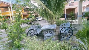 Un patio con un banco de piedra y dos ruedas de piedra, rodeado de plantas tropicales y una palmera.