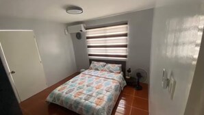 Un dormitorio con cama, ventana con persianas, aire acondicionado instalado en la pared y un ventilador.
