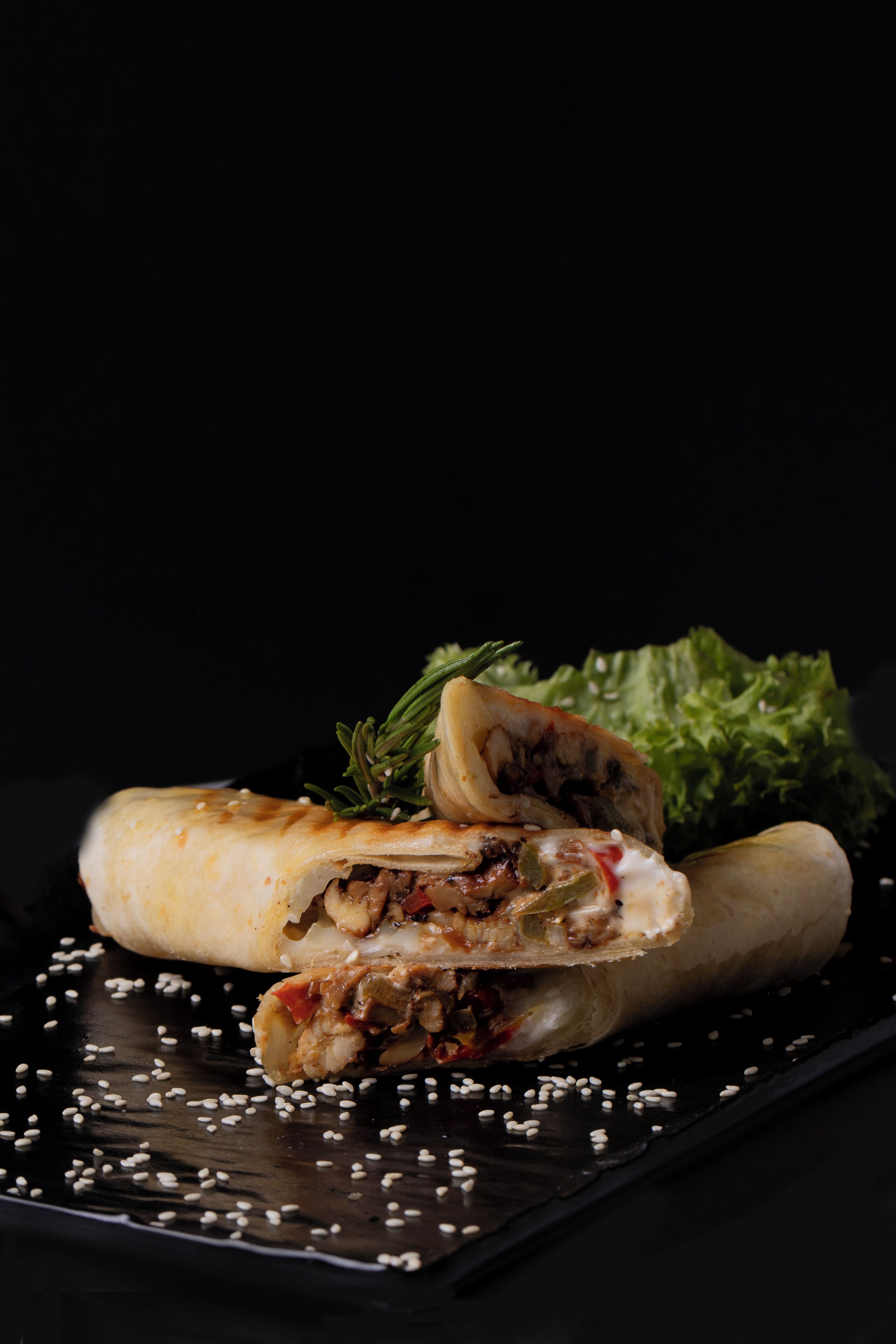 Un alimento enrollado con relleno visible, adornado con una ramita de romero, sobre un plato oscuro espolvoreado con semillas de sésamo.