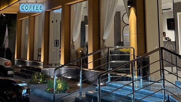 L’entrée d’un hôtel, avec un panneau lumineux au néon, un panneau indiquant qu’on y sert du café, et un escalier menant à l’entrée.