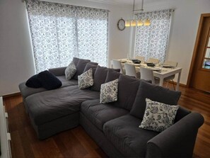 A modern living room with a grey sectional sofa, patterned cushions, a dining table with chairs, and a large window with patterned curtains. - Oceanview Gateway 
Beautiful 2Bed/2Bath Apartment (Caniço)