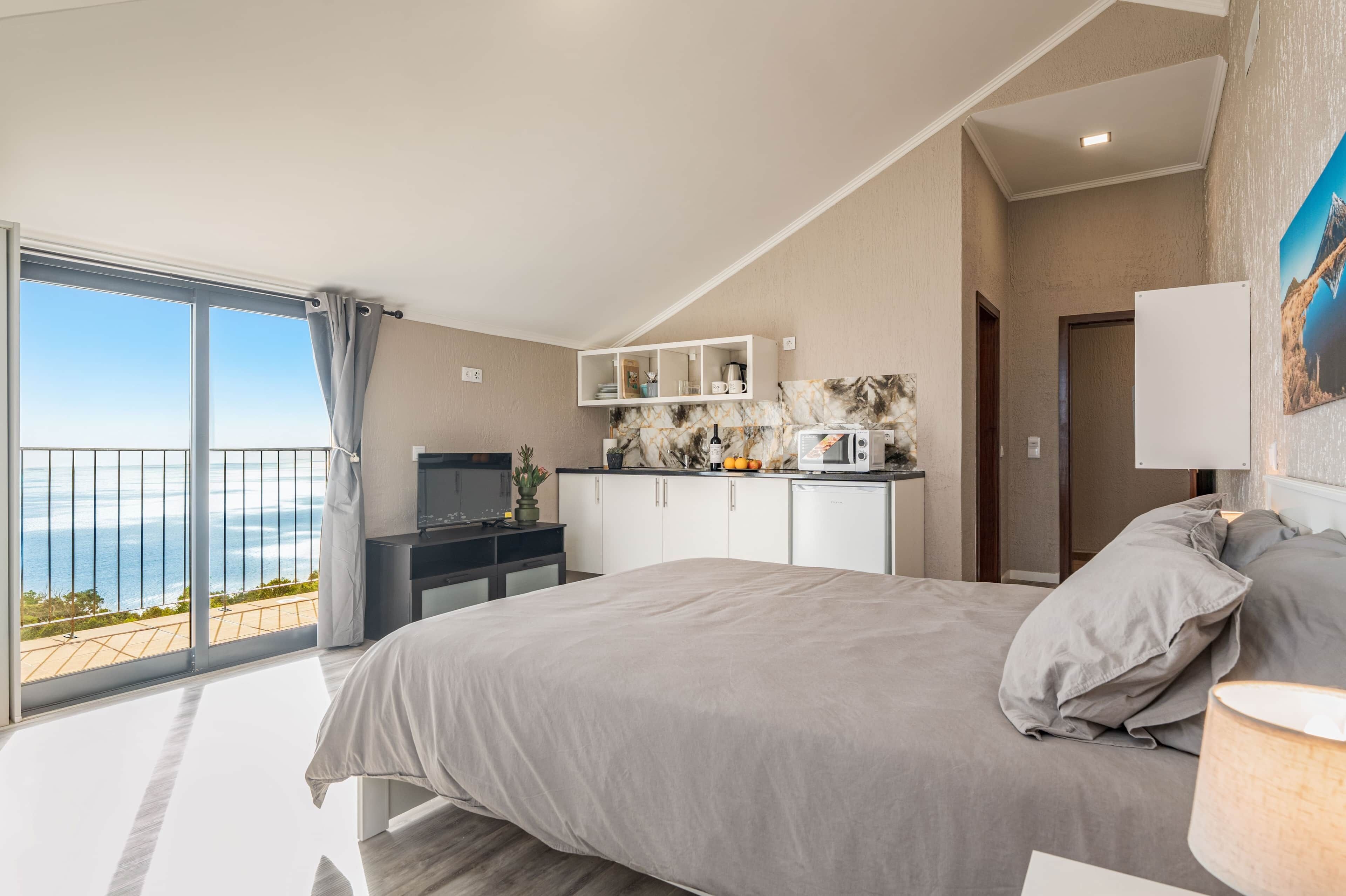 A bedroom with a bed, a TV, a kitchenette, and a balcony with a view of the sea.