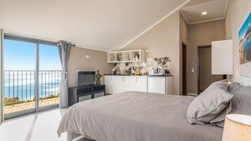A bedroom with a bed, a TV, a kitchenette, and a balcony with a view of the sea.