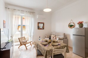 A dining area with a table set for two, a sofa, a refrigerator, and a window with curtains. - GuestHost - Comfortable 60 mq apartment, perfect for 4 people, located on the third floor of a building with lift.The property is located a few steps from the historic center of Sanremo, in an area full of services. In the immediate vicinity there is the (Sanremo)