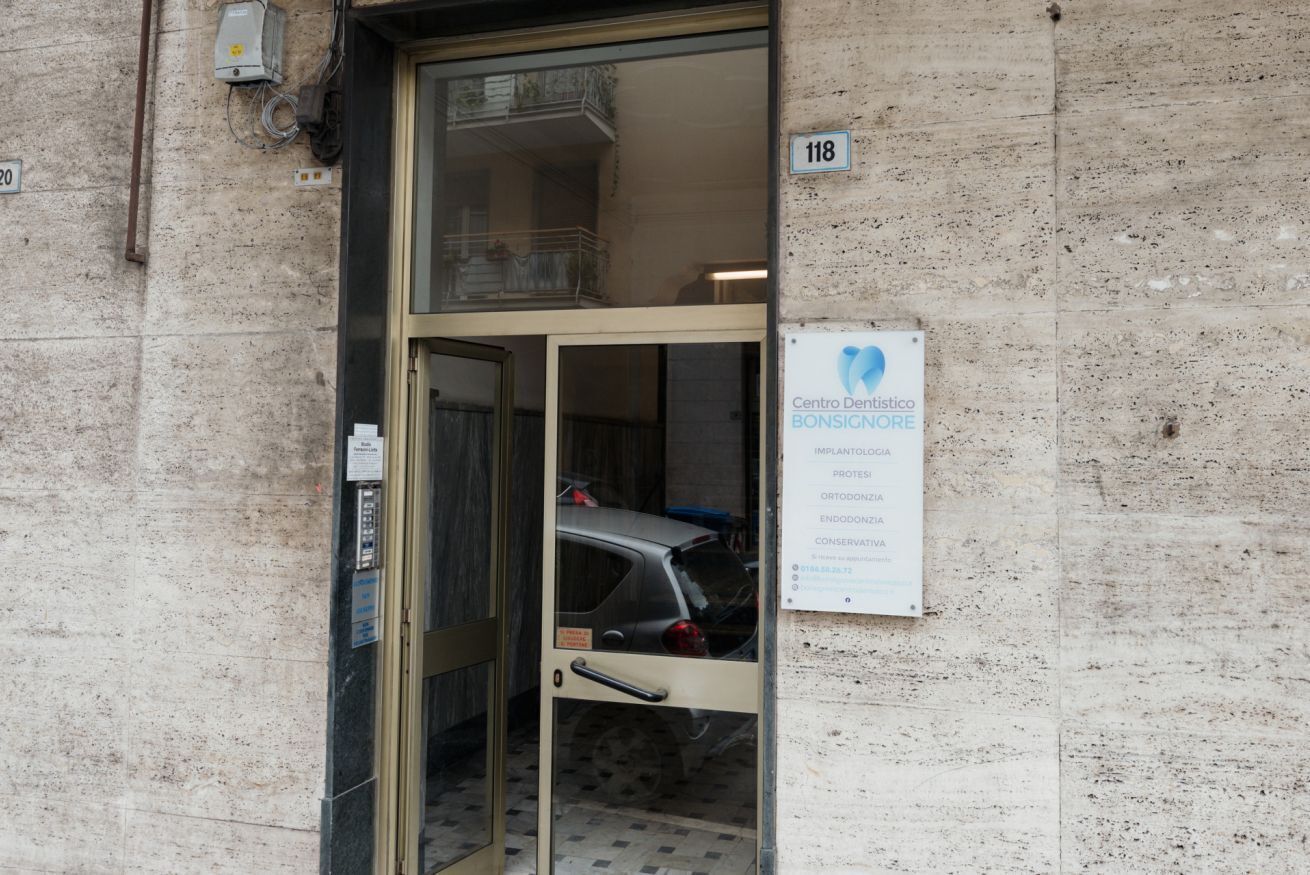 Een gevel met een glazen deur, een bord met de tekst 'Centro Dentistico Bonsignore' en een zichtbare auto.