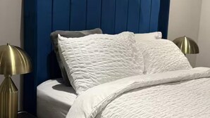 En seng med et blåt polstret sengegavl, hvide sengetøj og to gyldne lamper på hver side.