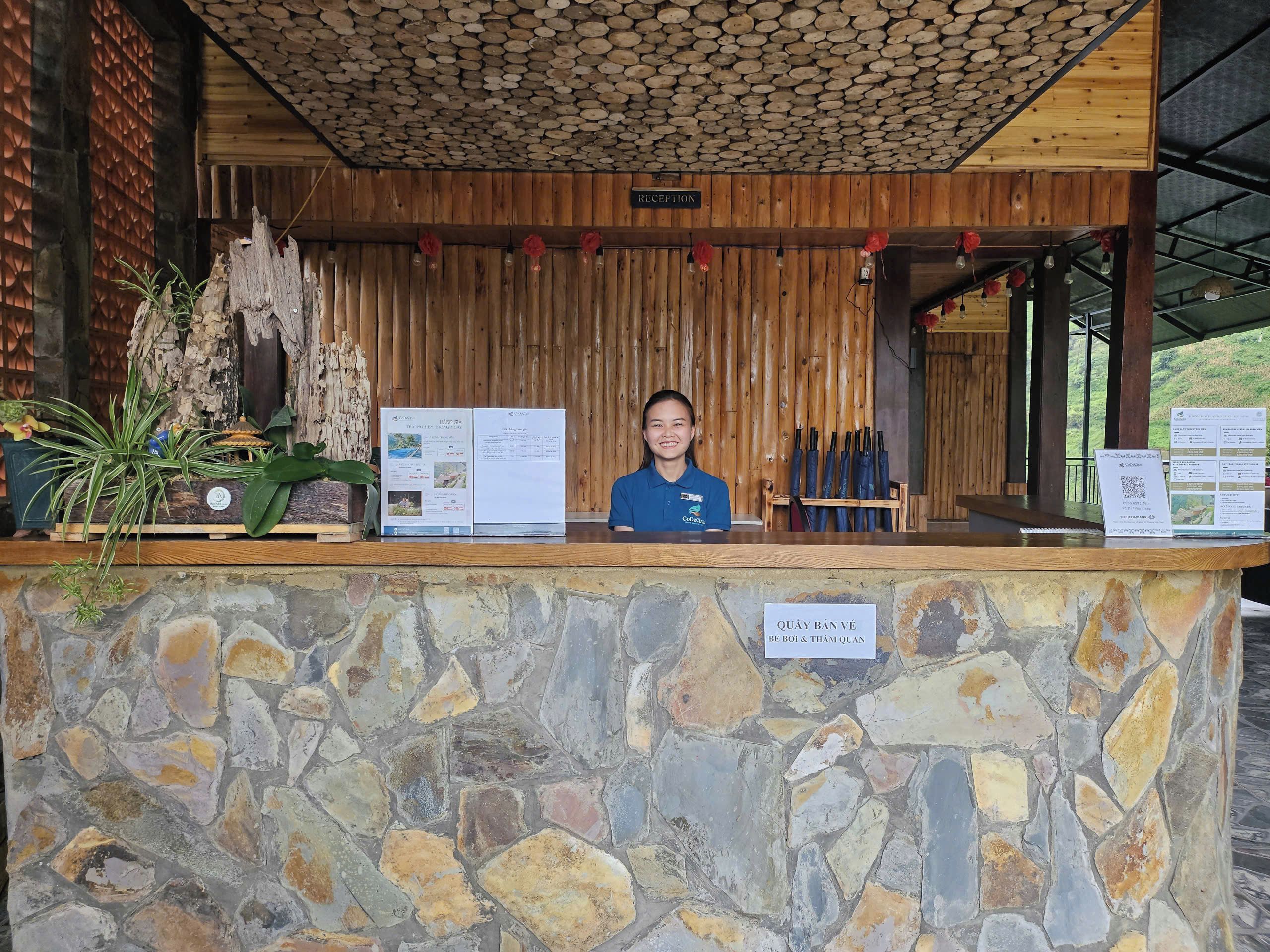 Una recepción con pared de piedra, paneles de madera y un cartel que indica 'QUÁY BÂN VÉ BE BỘI & THAM QUAN'.