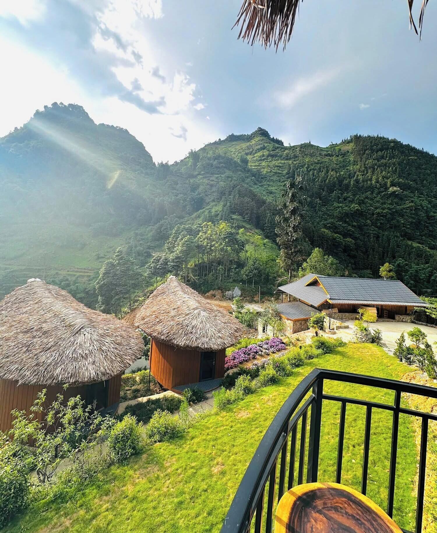 A rural setting with thatched-roof huts and a traditional building with a tiled roof, surrounded by lush greenery and hills.