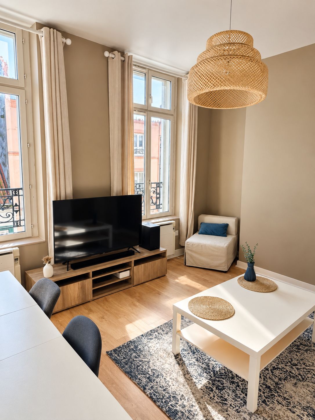 A modern living room with a flat-screen TV, a white dining table, and a woven pendant light.