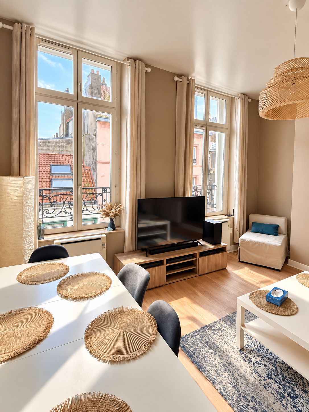 A modern living room with a flat-screen TV, a sofa, a dining table with placemats, and large windows with curtains.