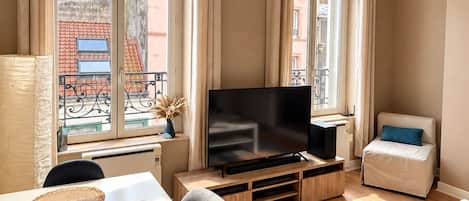 A modern living room with a flat-screen TV, a sofa, a dining table with placemats, and large windows with curtains.