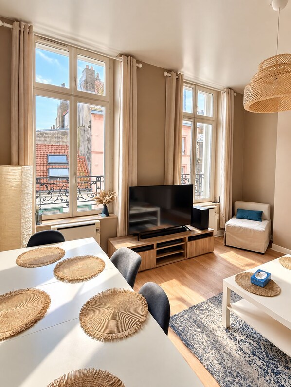 A modern living room with a flat-screen TV, a sofa, a dining table with placemats, and large windows with curtains.