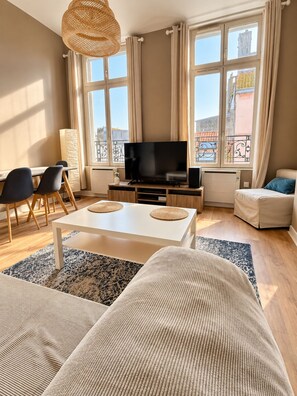 A modern living room with a flat-screen TV, a white coffee table, a sofa, and a dining area with chairs.